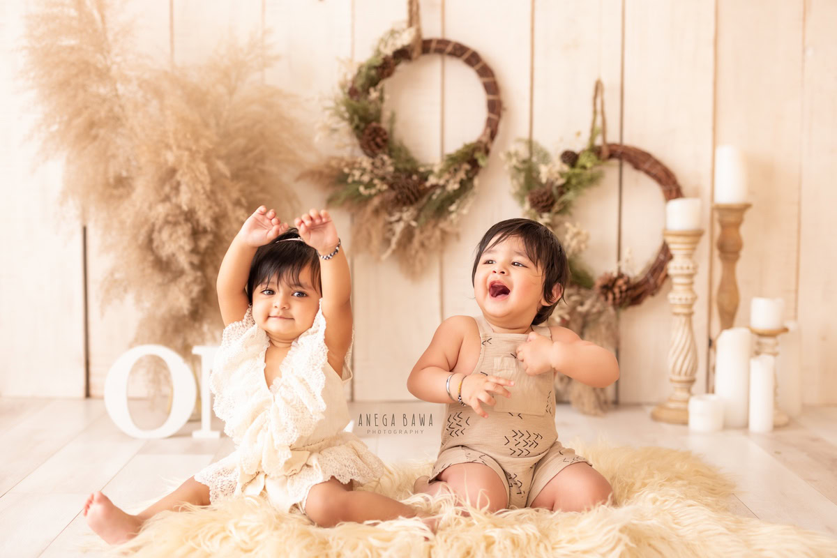 Capture the charm of a 1-year-old boy and girl sitting on a beige rug, surrounded by candle stands and leafy wooden frames on the wall. The bushes and beige backdrop create a perfect setting for this adorable sitter photography session in Delhi and Gurgaon by Anega Bawa.