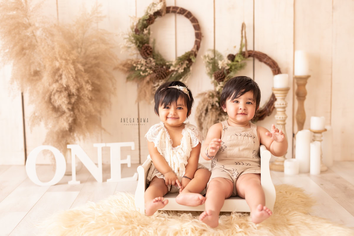 For a sitter photo shoot in Delhi and Gurgaon by Anega Bawa, a 1-year-old boy and girl are adorably seated in a wooden cot. The beige backdrop is beautifully complemented by leafy wooden frames, bushes, and a charming candle stand, creating a serene and natural setting. This exquisite setup captures the innocence and joy of childhood, preserving these precious moments in a timeless photograph.