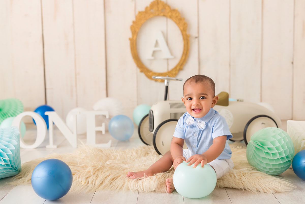 One-year-old boy holding a balloon and pompom on a beige rug with an alphabet frame on the wall against a beige backdrop, capturing the adorable moments of his pre-birthday photoshoot in Delhi by Anega Bawa, Gurgaon, Noida.