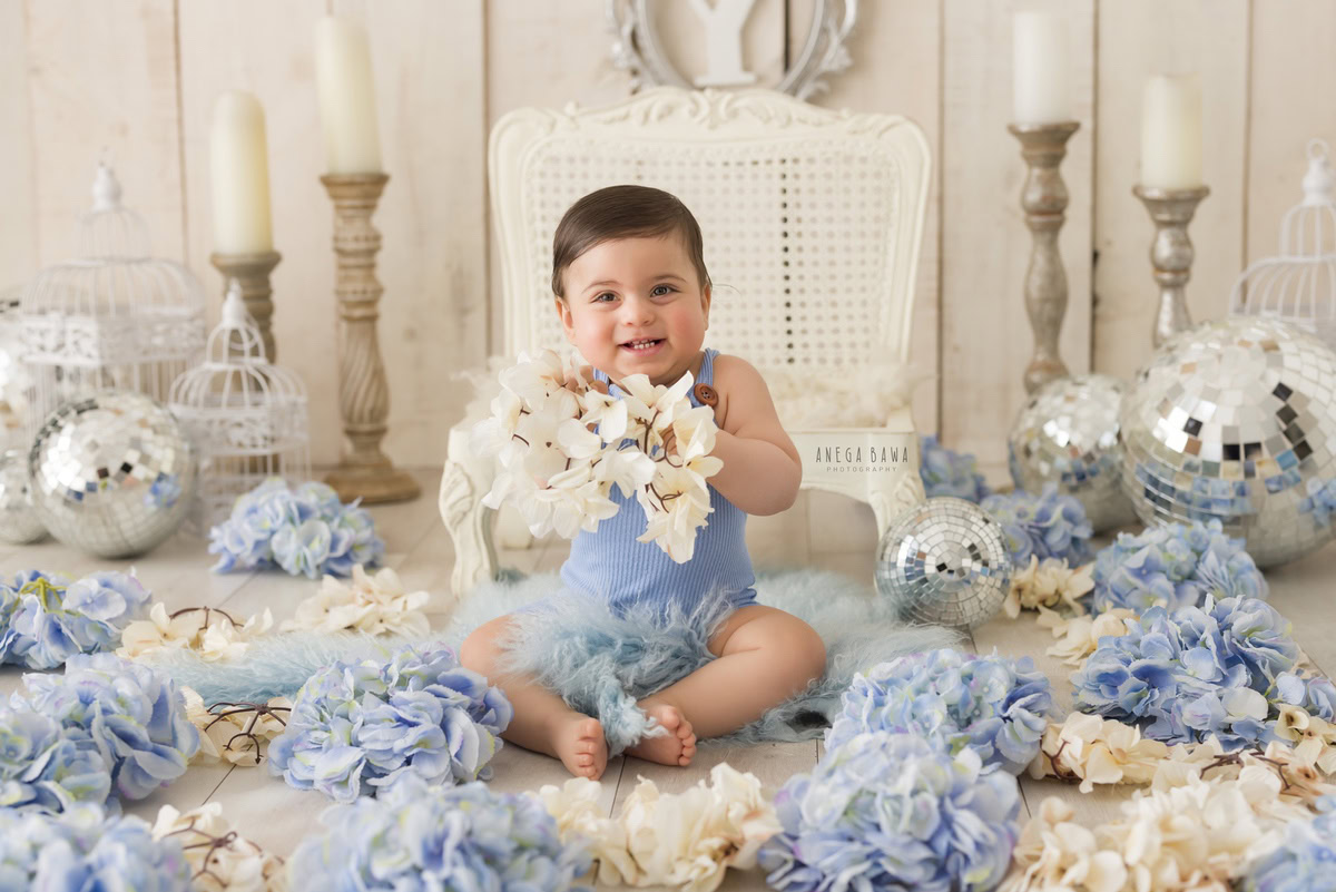 In this first birthday photoshoot by Anega Bawa in Delhi, a 1-year-old boy holds a flower while sitting on a white wooden chair. The backdrop features discoballs and a candle stand, creating a charming and festive scene. The photoshoot captures the boy's innocence and joy, marking a special milestone in a memorable setting.