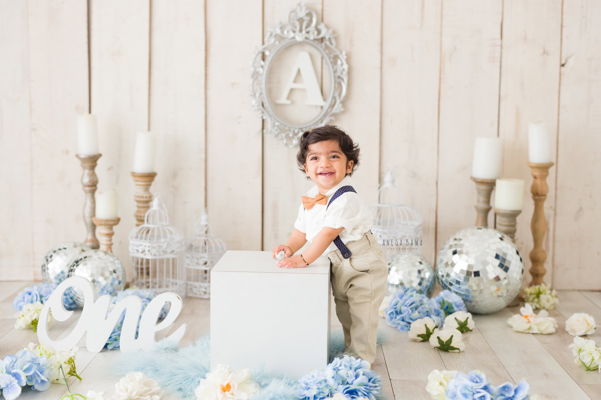 One-year-old boy holding a white stool amidst candle stands and a silver frame on the wall, with discoballs adorning the scene, on a blue rug surrounded by blue flowers against a pristine white backdrop, capturing the enchantment of his pre-birthday photoshoot in Delhi by Anega Bawa, Gurgaon, Noida.