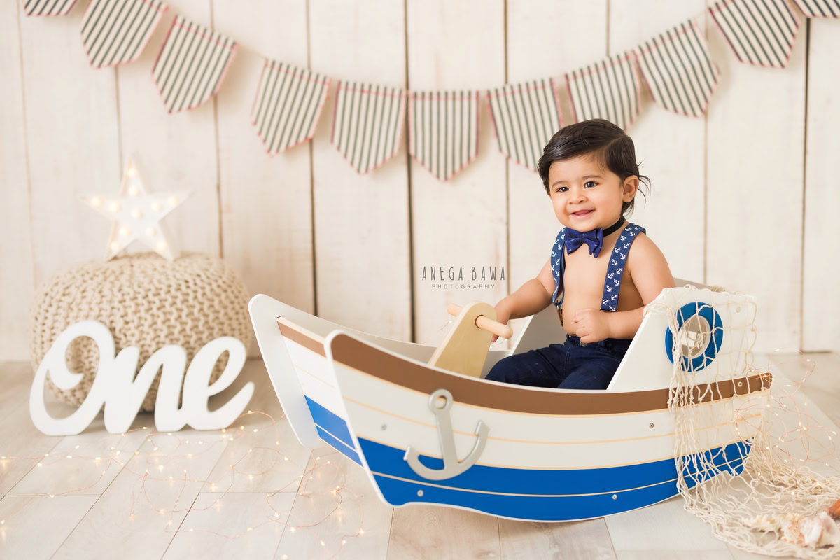 For a sitter photo shoot in Delhi and Gurgaon by Anega Bawa, a 1-year-old boy looks adorable in a toy spaceship with a blue bow against a beige backdrop. This charming setting captures the wonder and excitement of childhood, creating a timeless keepsake of these precious moments.