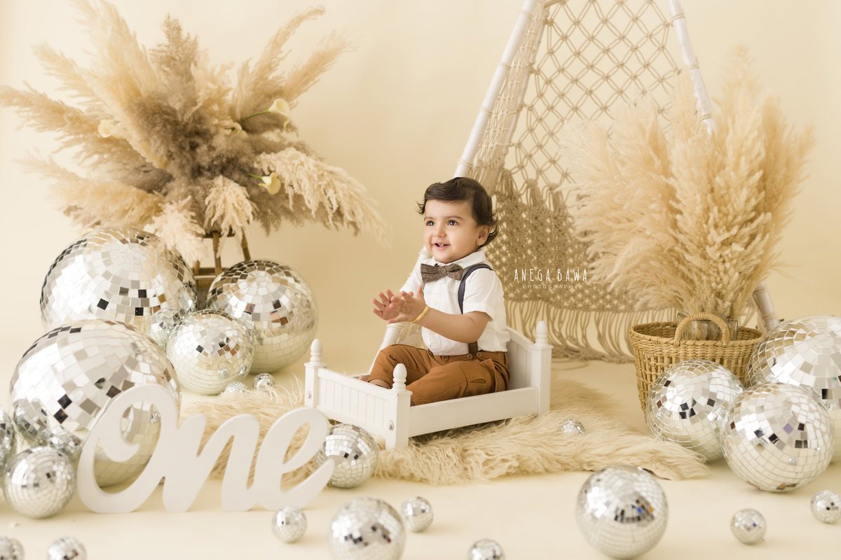Watch your little one bask in the comfort of a white cot, surrounded by the playful allure of disco balls and verdant bushes against a serene beige backdrop. Capture these heartwarming moments with Anega Bawa Photography's sitter photography services in Delhi and Gurgaon.