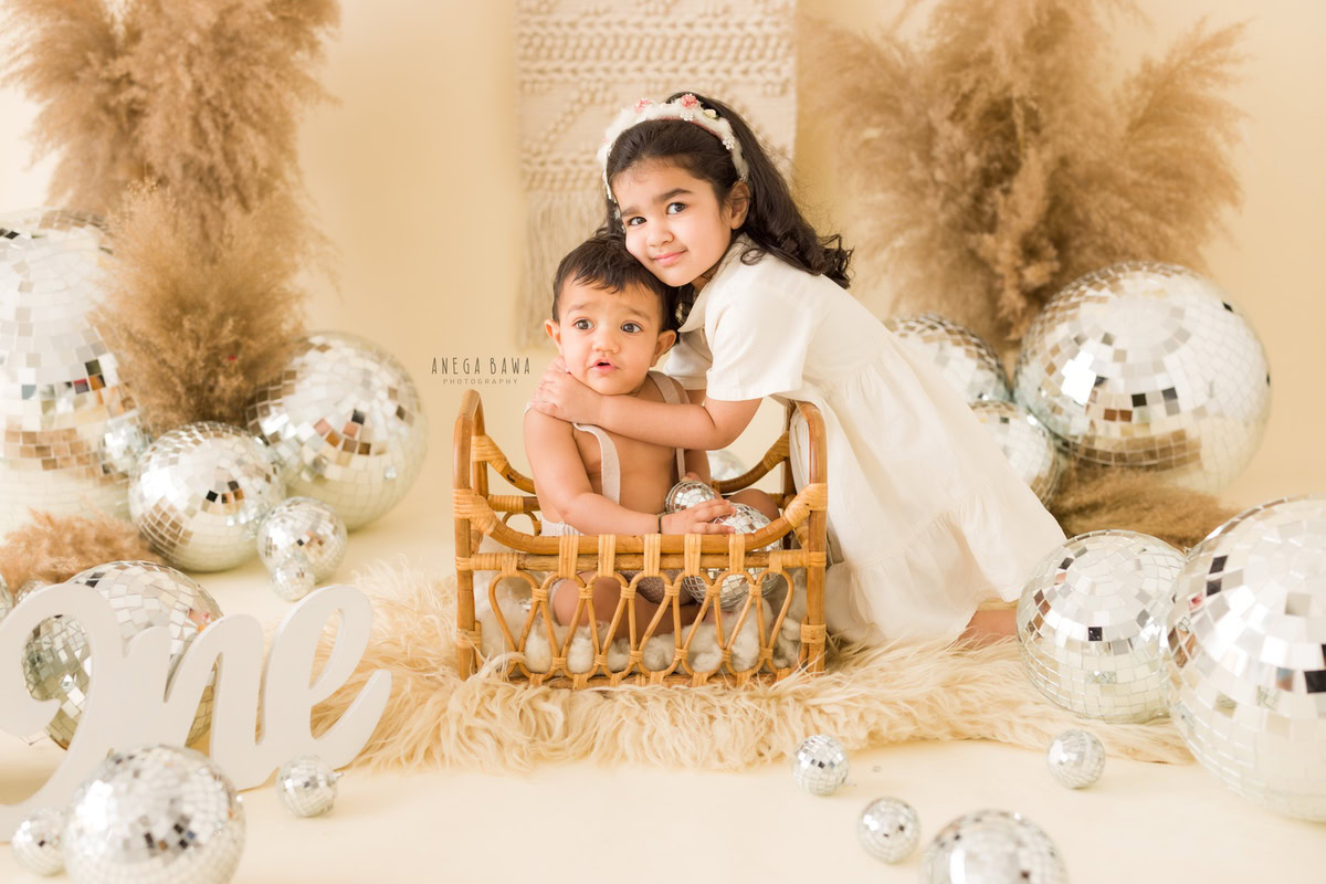 Celebrate the bond between your little ones as they share giggles and playful moments in a wooden cot, amidst brown bushes and sparkling disco balls against a calming beige backdrop. Let Anega Bawa Photography in Delhi and Gurgaon immortalize these precious sibling memories with their sitter photography services.