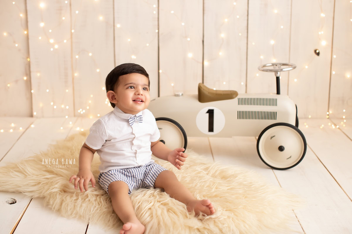 For a sitter photo shoot in Delhi and Gurgaon by Anega Bawa, a 1-year-old boy is captured in an enchanting setting. Seated on a toy scooter on a beige rug, surrounded by fairy lights, this delightful scene beautifully captures the innocence and charm of the milestone moment.