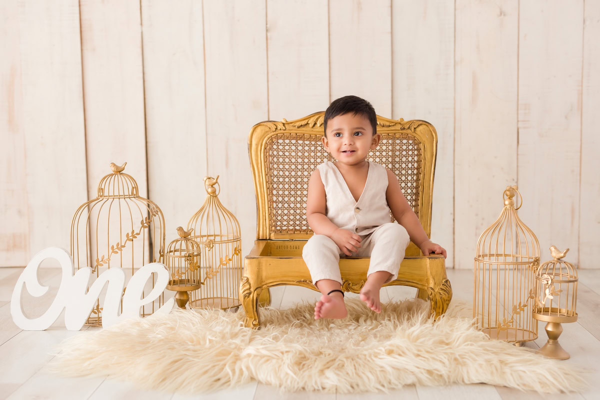Captivating one-year-old boy perched on a regal golden chair, atop a luxurious beige rug, surrounded by golden castles against a stunning beige backdrop, showcasing his enchanting first birthday photoshoot in Delhi, by Anega Bawa, Gurgaon, Noida