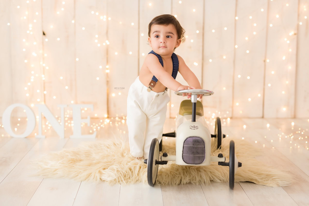 One-year-old boy on a toy scooter on a beige rug with fairy lights against a beige backdrop, capturing the magical ambiance of his pre-birthday photoshoot in Delhi by Anega Bawa, Gurgaon, Noida.