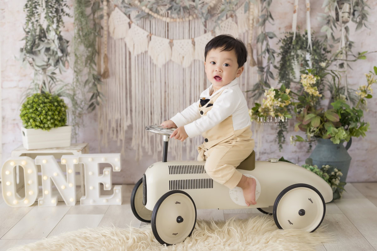 Anega Bawa's toddler photoshoot in Delhi, Gurgaon, and Noida features a 1-year-old boy on a toy scooter. The setup includes a white backdrop, a green pot, and a leafy fringe, creating a charming and vibrant scene perfect for capturing precious moments.