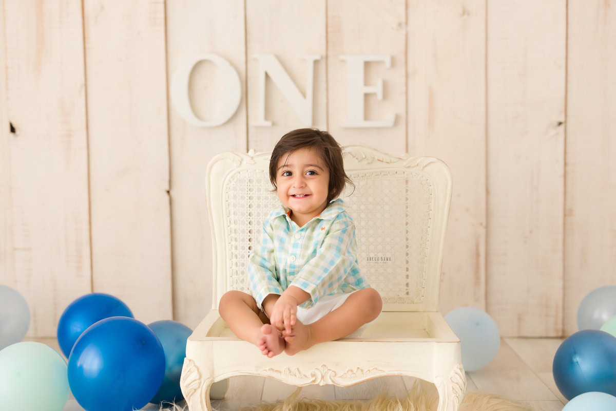 Capturing the delightful essence of a pre-birthday photoshoot in Delhi, witness a charming one-year-old boy seated on a pristine white chair against a captivating beige backdrop adorned with whimsical balloons. Photographed by the talented Anega Bawa in Gurgaon, Noida