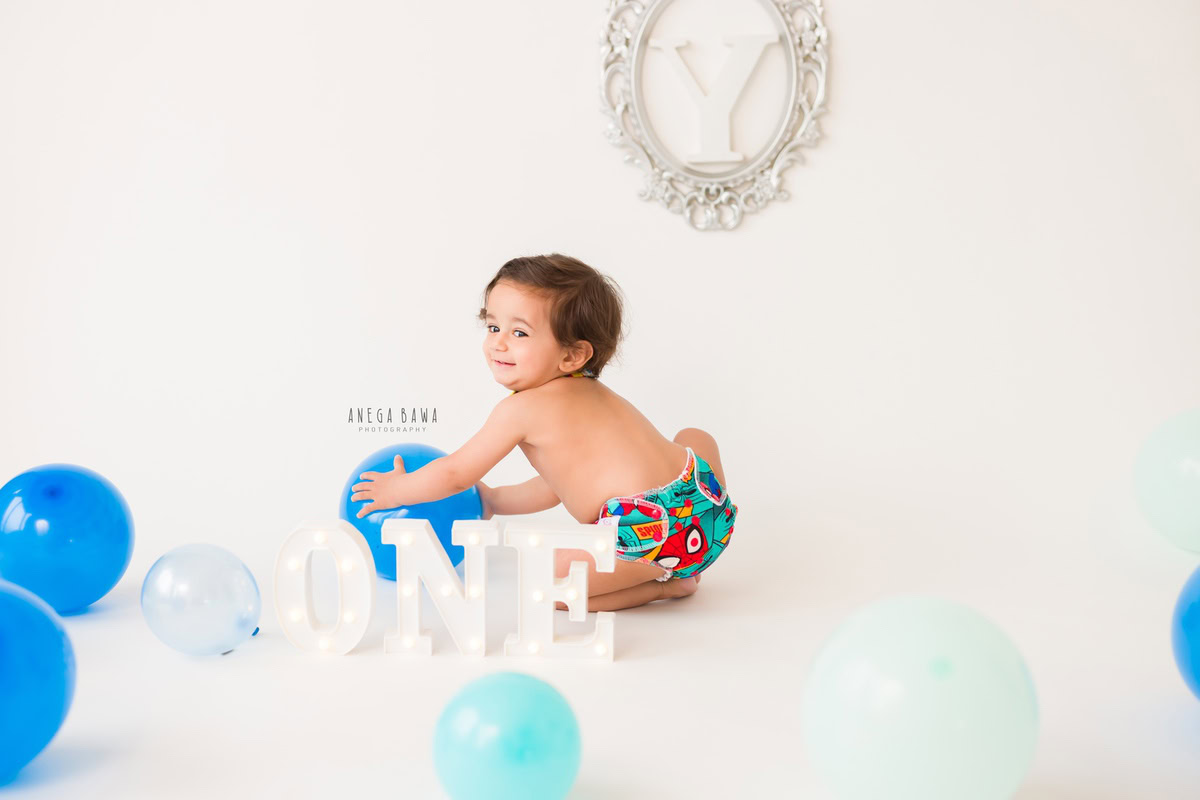 For a sitter photo shoot in Delhi and Gurgaon by Anega Bawa, a 1-year-old boy is joyfully playing with a blue balloon against a pristine white backdrop. The scene is adorned with an alphabet frame on the wall, creating a charming and playful ambiance that perfectly captures this precious moment.