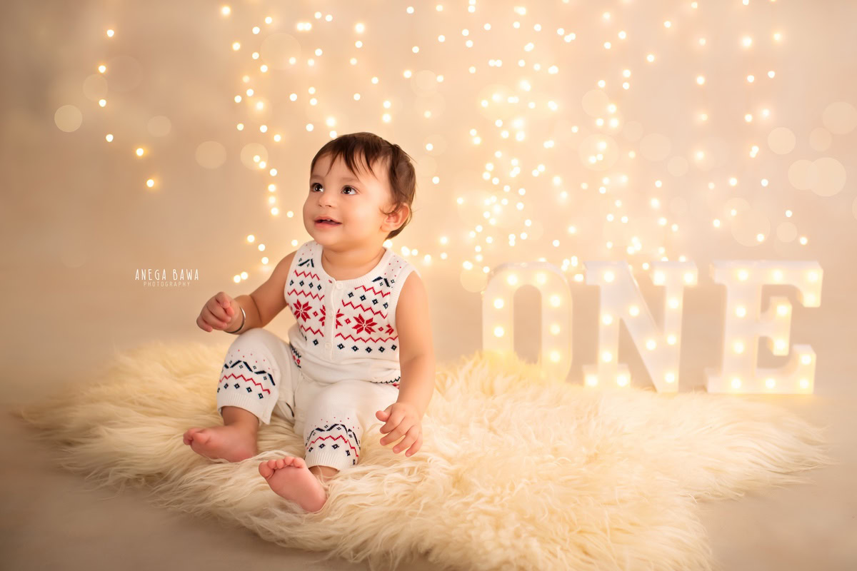 Cherubic one-year-old boy seated on a cozy beige rug, illuminated by twinkling fairy lights, capturing the magic of his first birthday photoshoot in Delhi, by Anega Bawa, Gurgaon, Noida