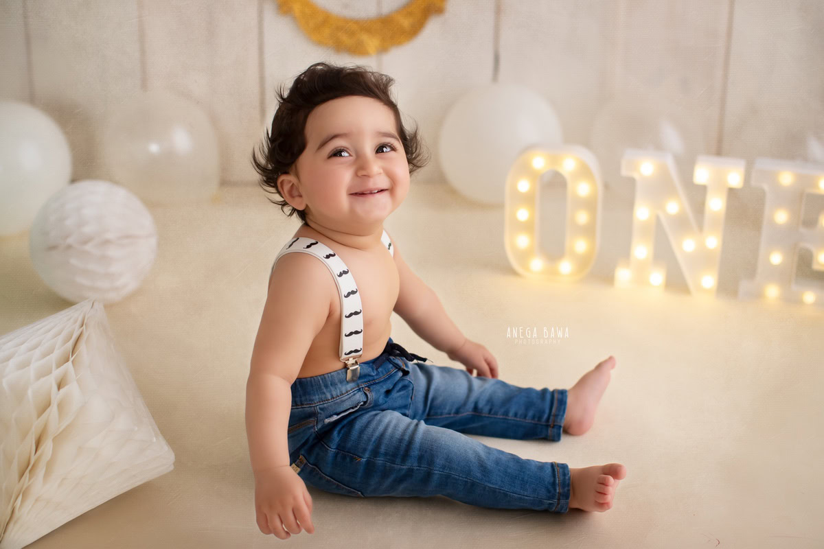 Capture the joy of your 1-year-old boy with a sitter photoshoot by Anega Bawa Photography in Delhi and Gurgaon. Your little one sits on the floor against a beige backdrop, surrounded by colorful balloons, creating a delightful and memorable scene.