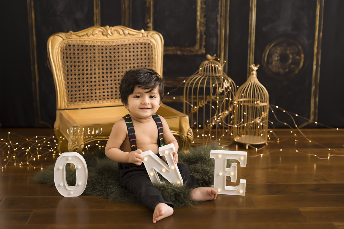 Radiant one-year-old boy seated on the floor beside a majestic golden chair adorned with golden castles, bathed in the gentle glow of fairy lights, commemorating his enchanting first birthday photoshoot in Delhi, by Anega Bawa, Gurgaon, Noida