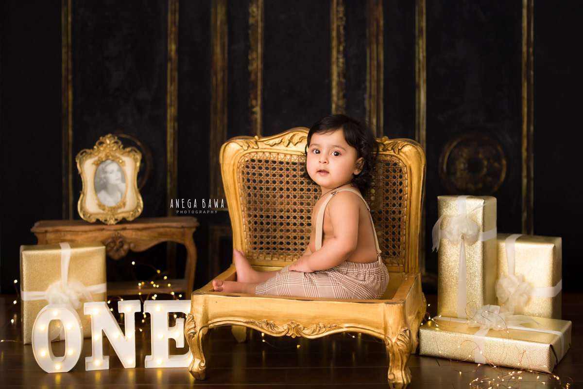 Capture the magical moment of a 1-year-old boy seated on a golden chair, surrounded by gifts, a golden photo frame, and fairy lights. This enchanting setup is perfect for a sitter photography session in Delhi and Gurgaon by Anega Bawa.