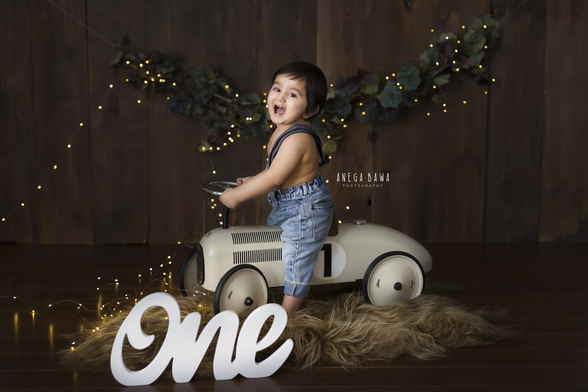 Capture the adorable charm of a 1-year-old boy seated on a toy scooter, set against a brown rug and backdrop adorned with golden lights. Ideal for a sitter photography session in Delhi and Gurgaon by Anega Bawa.