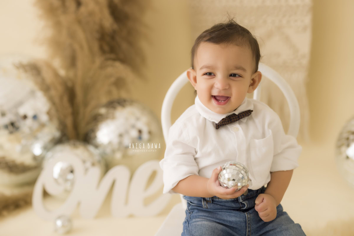 Captivating one-year-old boy seated on a pristine white chair, holding a sparkling disco ball amidst rustic brown bushes, against a serene beige backdrop, immortalizing his memorable first birthday photoshoot in Delhi, by Anega Bawa, Gurgaon, Noida