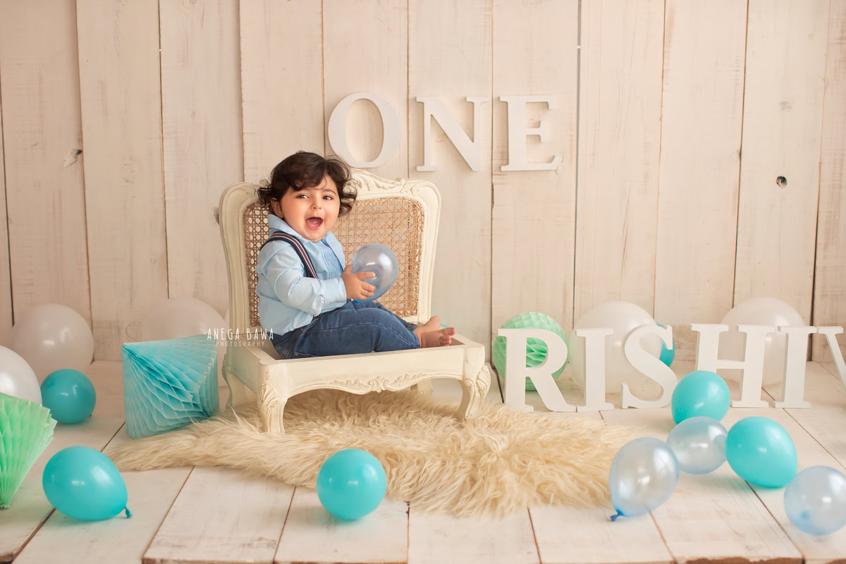 One-year-old boy elegantly seated on a rustic wooden chair amidst a backdrop adorned with vibrant balloons, set against a serene beige background, capturing the magic of his pre-birthday photoshoot in Delhi by the talented Anega Bawa in Gurgaon, Noida.