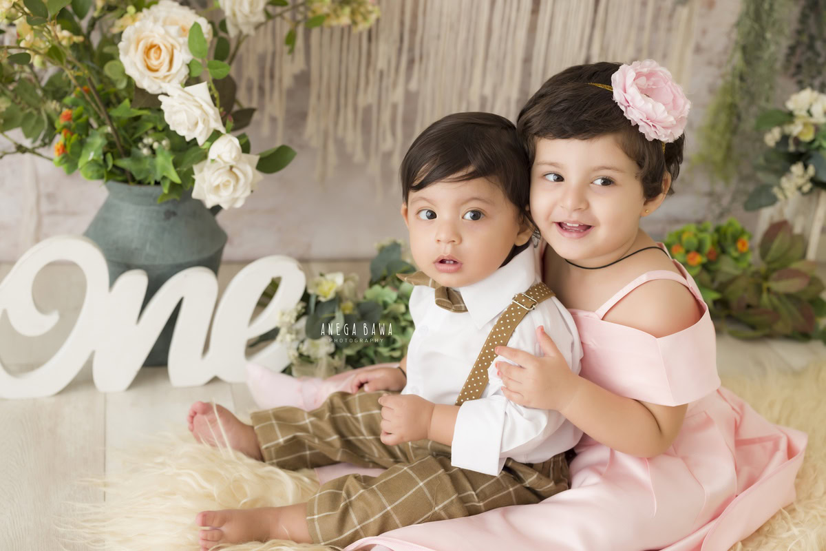 For a captivating toddler photoshoot in Delhi, Gurgaon, and Noida by Anega Bawa, the setup features a 1-year-old boy with his sibling, surrounded by green pots on a beige rug against a beige backdrop. This serene and harmonious setting is perfect for capturing precious sibling moments.