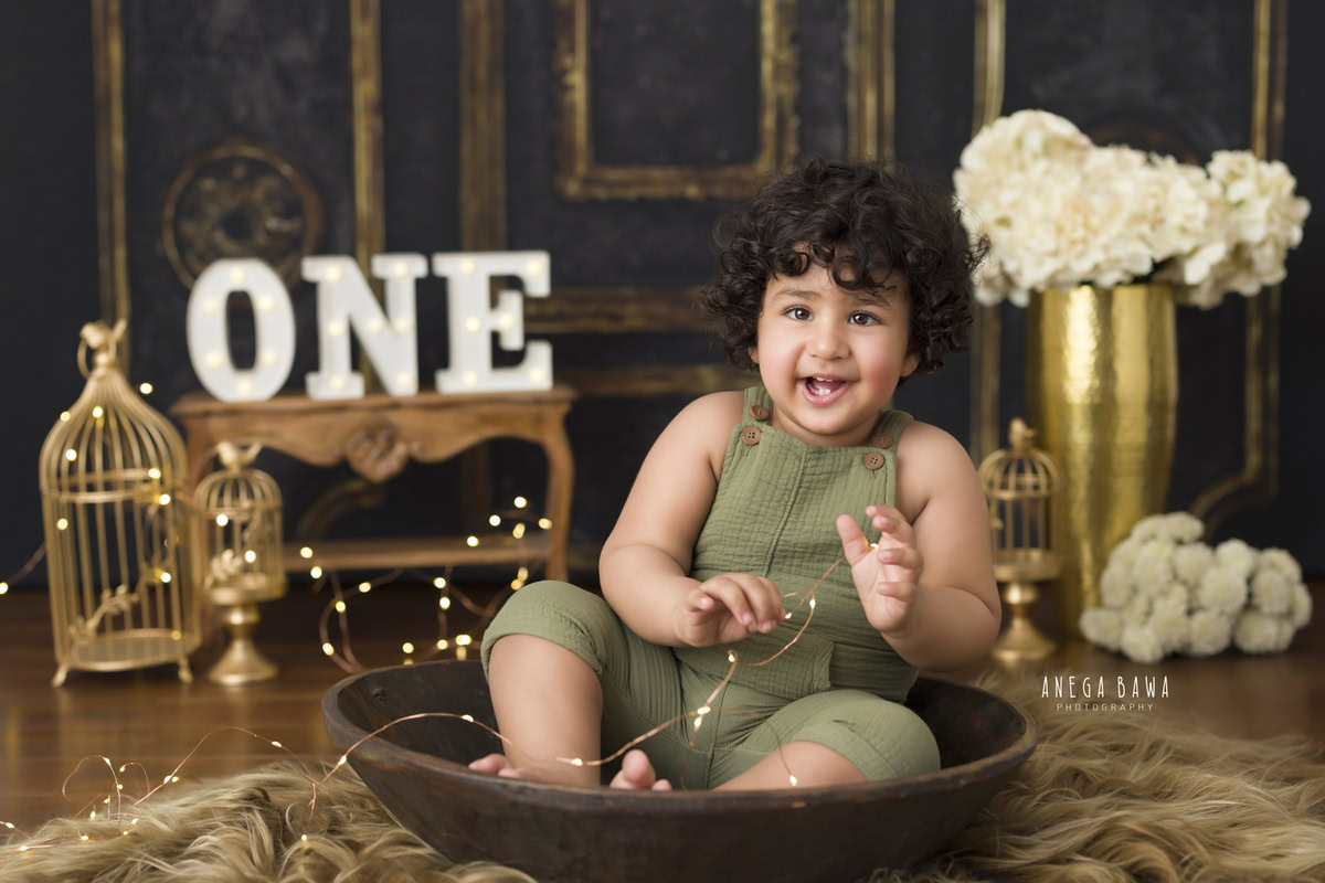 Capture precious moments of your 1-year-old boy with a sitter photoshoot by Anega Bawa Photography in Delhi and Gurgaon. Dressed in a military green dungaree, your little one sits in a brown tub, surrounded by golden castles and white flowers, creating a magical and memorable scene.