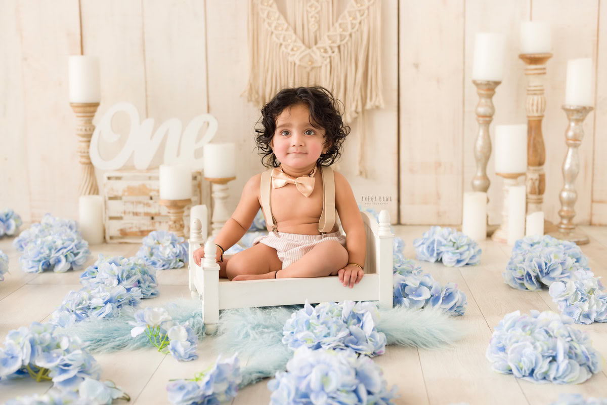 Adorable one-year-old boy nestled in a pristine white cot, surrounded by the soft glow of candle stands against a sophisticated beige backdrop, in a charming pre-birthday photoshoot captured by Anega Bawa in the heart of Delhi, Gurgaon, Noida.