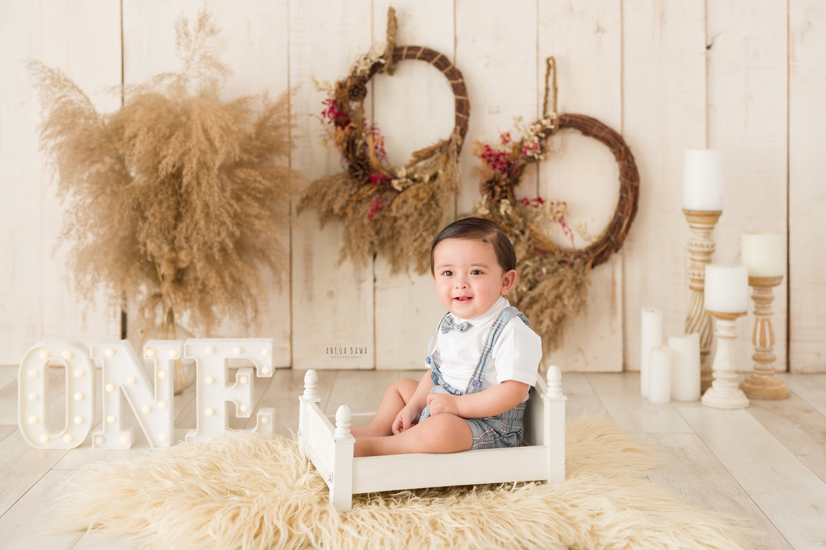 Delight in the sight of your 1-year-old boy seated in a white cot, surrounded by the warm glow of candlelight and wooden floral bushes, with a charming frame adorning the wall against a soothing beige backdrop. Trust Anega Bawa Photography in Delhi and Gurgaon to capture this beautiful moment with their impeccable sitter photography services.