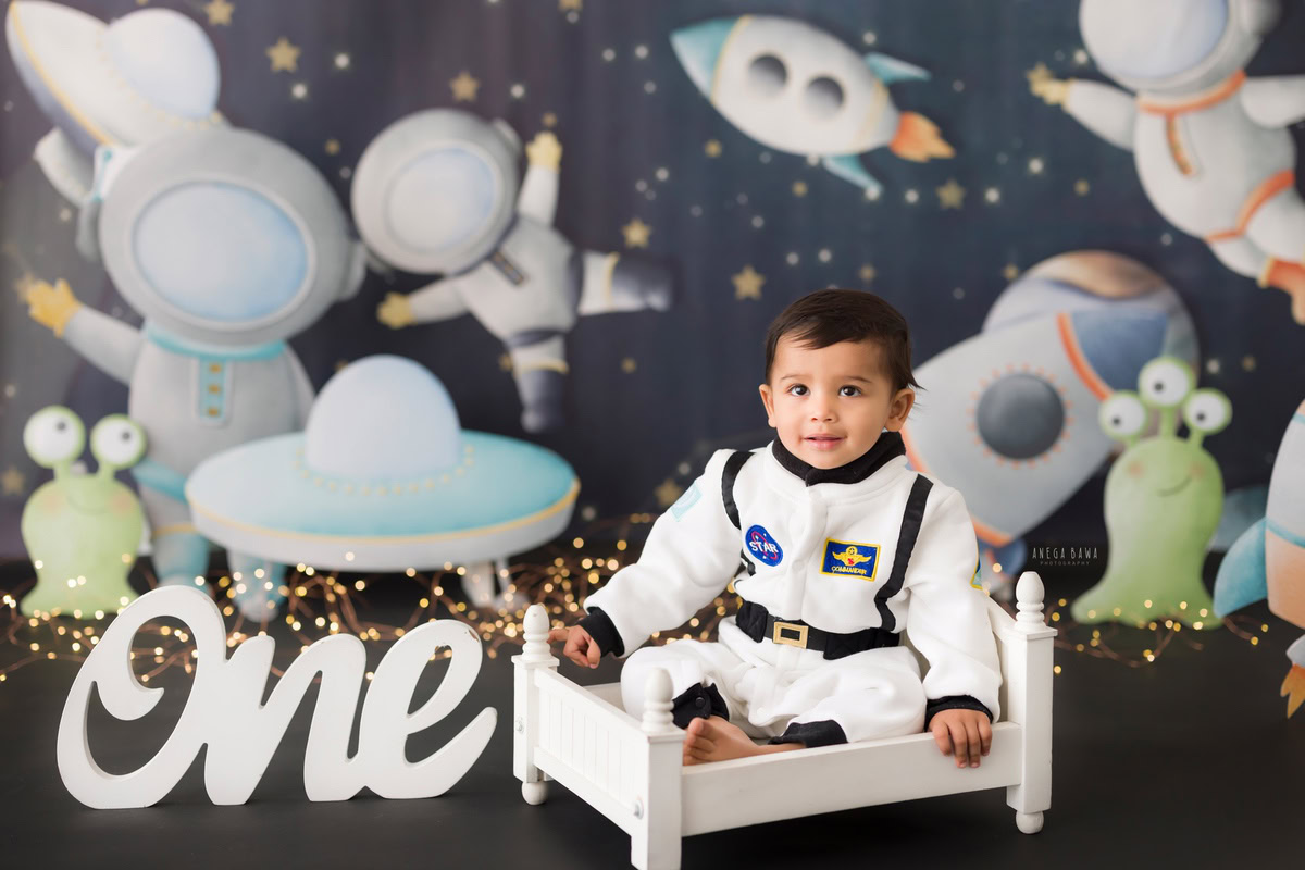 For a delightful toddler photoshoot in Delhi, Gurgaon, and Noida by Anega Bawa, we feature a 1-year-old boy sitting in a white cot against a space-themed backdrop. This creative setting captures the wonder and imagination of childhood perfectly.