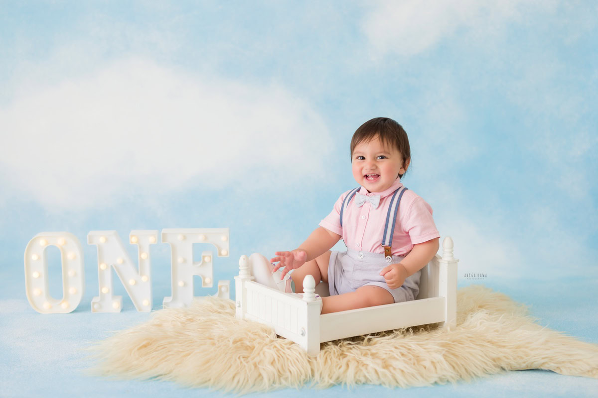 For a captivating toddler photoshoot in Delhi, Gurgaon, and Noida by Anega Bawa, we present a charming 1-year-old boy sitting in a white cot on a beige rug against a dreamy cloudy backdrop. This enchanting setup beautifully captures the innocence and wonder of early childhood.