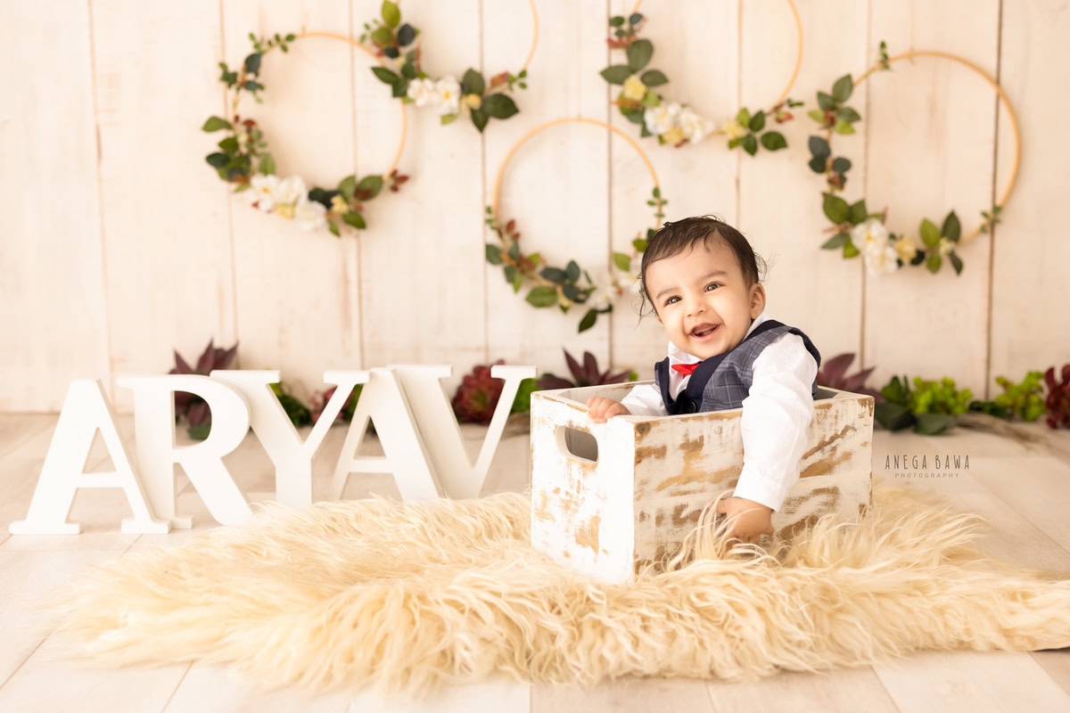 Capture the charm of your 1-year-old boy in a sitter photoshoot by Anega Bawa Photography in Delhi and Gurgaon. Your little one sits in a wooden basket on a beige rug, framed by beautiful floral circle frames on the wall, creating an adorable and timeless scene.
