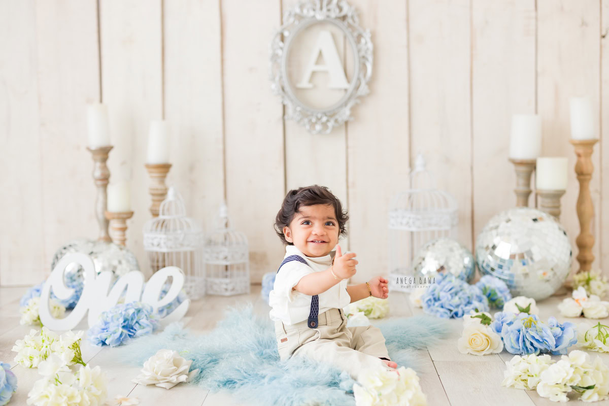 Immerse yourself in the adorable sight of your 1-year-old boy seated on a cozy blue rug, surrounded by shimmering discoballs and the soft glow of candlelight. Against a serene beige backdrop adorned with blue and white flowers, a silver frame on the wall adds a touch of elegance to the scene. Let Anega Bawa Photography in Delhi and Gurgaon capture this precious moment with their expert sitter photography services.