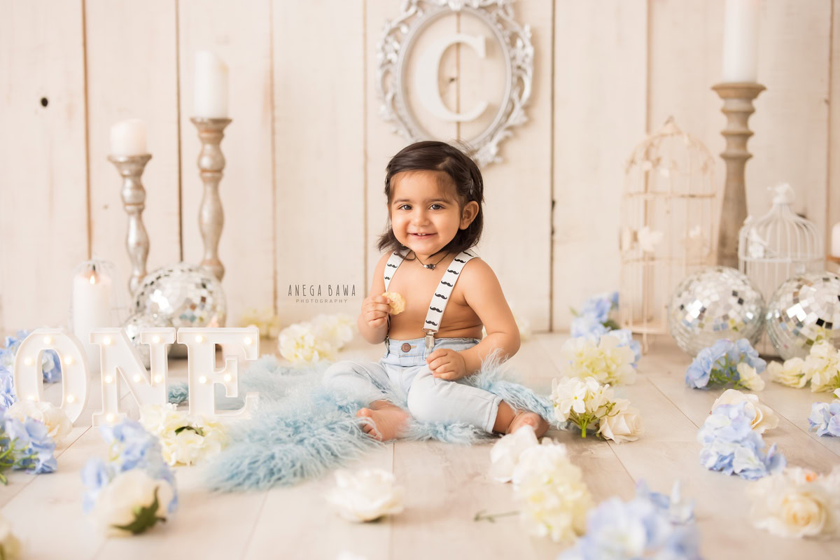 For a sitter photo shoot in Delhi and Gurgaon by Anega Bawa, a 1-year-old boy is sitting on a blue rug surrounded by disco balls and candle stands. The backdrop features a beige tone with an alphabet frame on the wall, creating a captivating and delightful setting for this memorable moment.