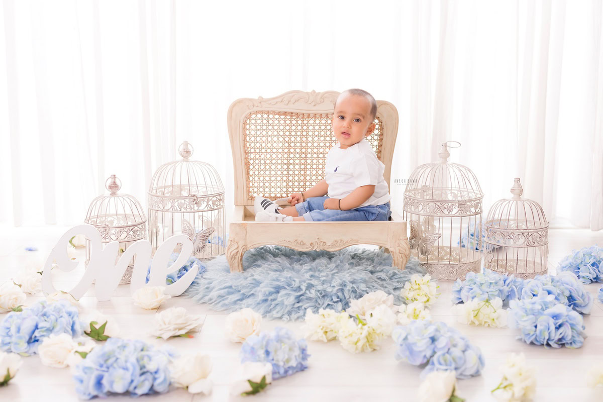 Adorable one-year-old boy sitting on a chair amidst a scattering of blue and white flowers, surrounded by elegant white castles against a pristine white backdrop, capturing the essence of his first birthday photoshoot in Delhi, by Anega Bawa, Gurgaon, Noida