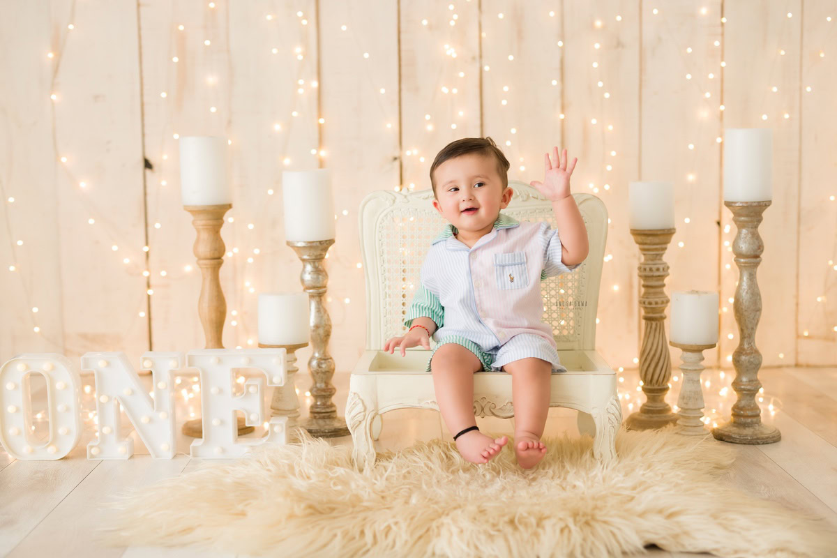 In a captivating pre-birthday photoshoot by Anega Bawa in Delhi, witness a cherubic one-year-old boy seated on a chair amidst the gentle glow of candle stands and fairy lights against a chic beige backdrop in Gurgaon, Noida.