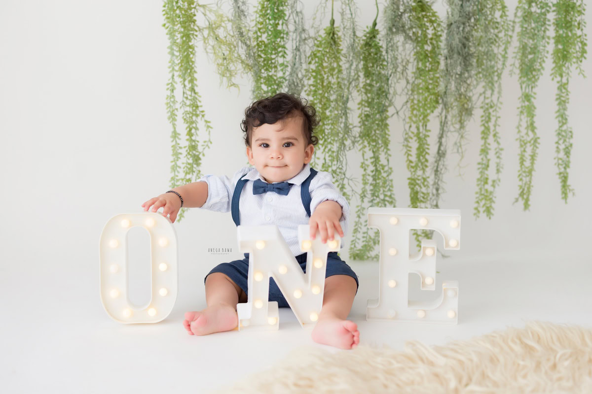 Experience the serenity of a baby's pre-birthday photoshoot by Anega Bawa in Delhi as a charming one-year-old boy sits gracefully on a cozy beige rug, surrounded by a lush leafy fringe, all against a pristine white backdrop in Gurgaon, Noida.