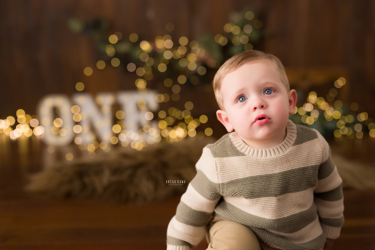 Immerse yourself in the enchanting ambiance of a baby's pre-birthday photoshoot in Delhi, where a delightful one-year-old boy sits on a warm brown rug, enveloped by the soft glow of fairy lights against a rich brown backdrop, expertly captured by Anega Bawa in Gurgaon, Noida.