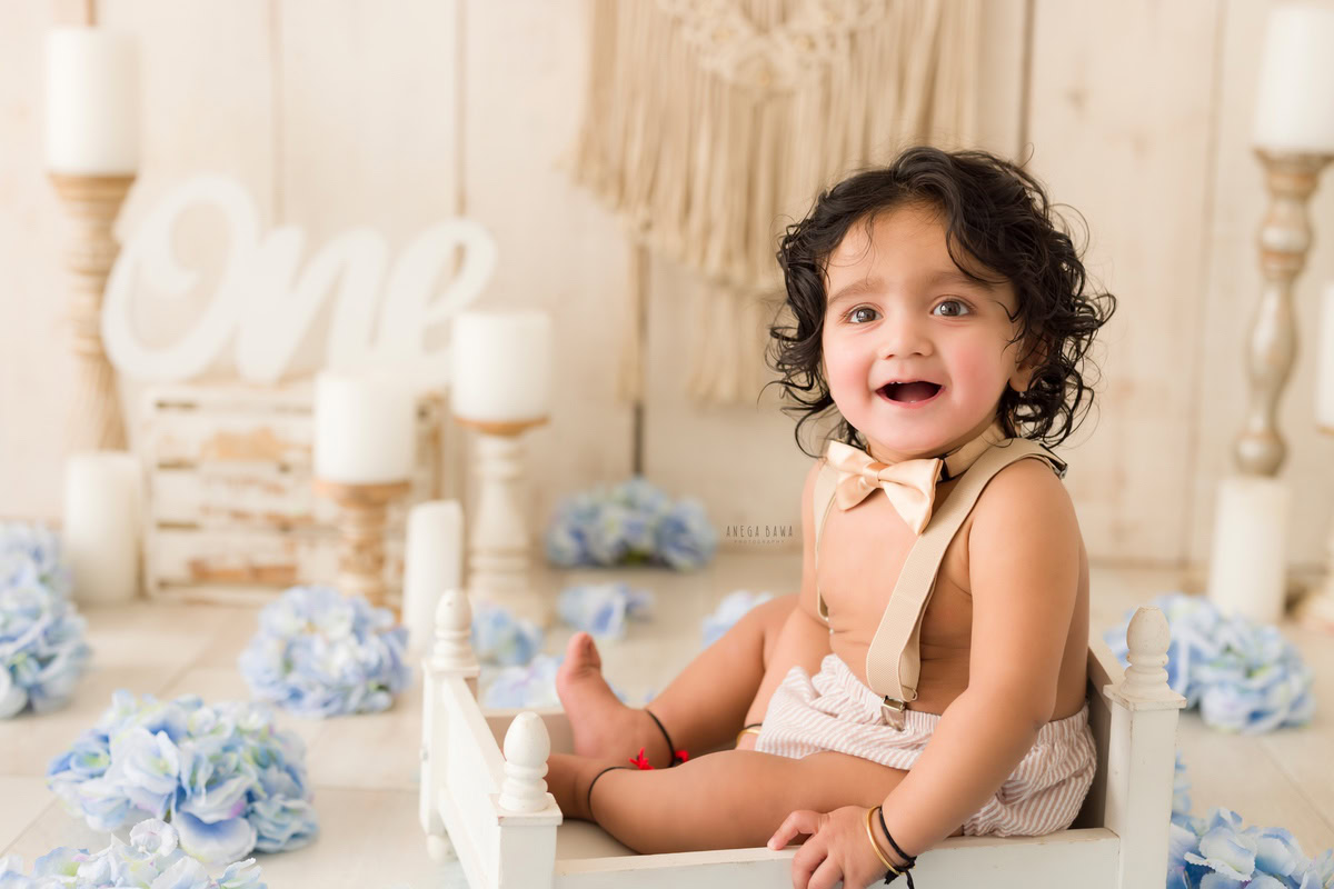 Step into the captivating world of a baby's pre-birthday photoshoot in Delhi, where a charming one-year-old boy sits amidst the soft glow of candle stands and delicate blue flowers against a serene beige backdrop, skillfully captured by Anega Bawa in Gurgaon, Noida.