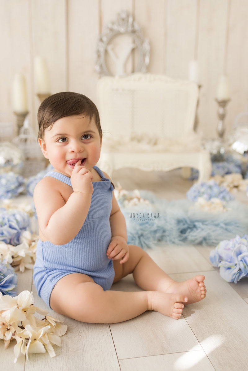 Embark on a journey into the whimsical world of a baby's pre-birthday photoshoot in Delhi, as a delightful one-year-old boy sits amidst the elegance of candle stands and delicate flowers against a pristine white backdrop, expertly captured by Anega Bawa in Gurgaon, Noida.