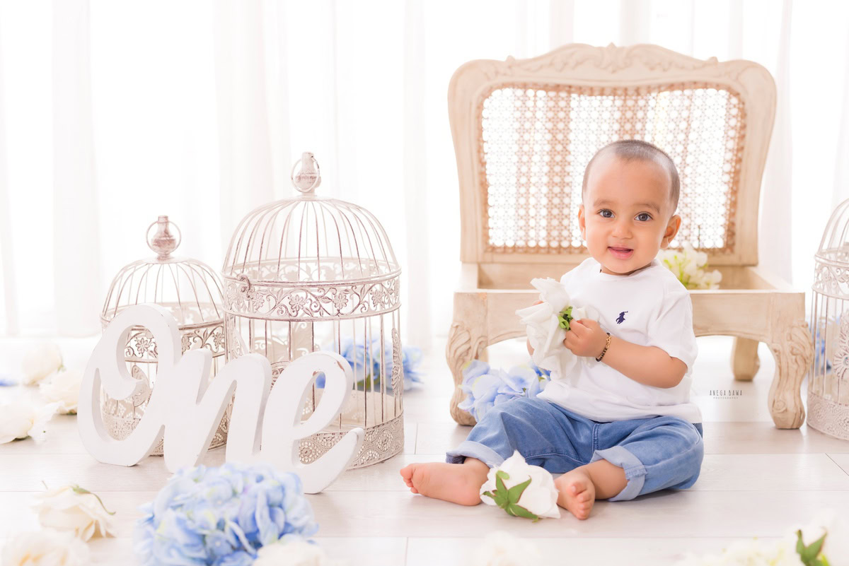 Sweet one-year-old boy sitting on the floor, holding a delicate flower, surrounded by charming white castles against a serene white backdrop, encapsulating the joy of his first birthday photoshoot in Delhi, by Anega Bawa, Gurgaon, Noida
