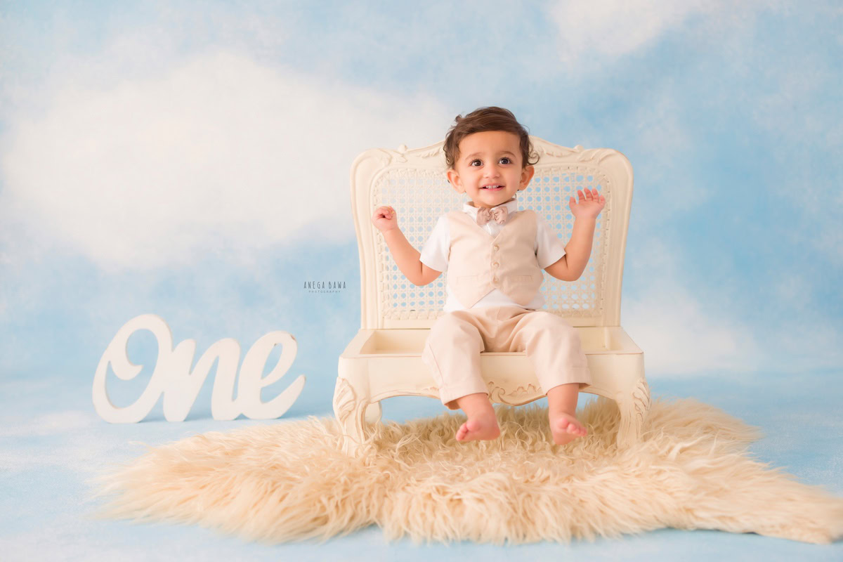 Delve into the enchanting atmosphere of a baby's pre-birthday photoshoot in Delhi, where a delightful one-year-old boy is seated on an elegant ivory chair, set against a dreamy cloudy backdrop and a cozy beige rug, skillfully photographed by Anega Bawa in Gurgaon, Noida.