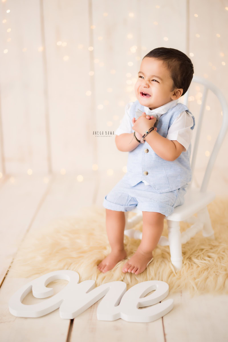 Captured by Anega Bawa, a 1-year-old boy sits on a white chair atop a beige rug against a beige backdrop adorned with golden lights. This enchanting scene, ideal for a sitter photo shoot in Gurgaon and Delhi, emanates warmth and charm.
