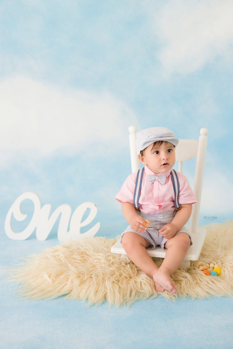 Captivating one-year-old boy sitting on a pristine white chair atop a cozy beige rug, set against a dreamy cloudy backdrop, crowned with a charming cap, commemorating his first birthday photoshoot in Delhi, by Anega Bawa, Gurgaon, Noida