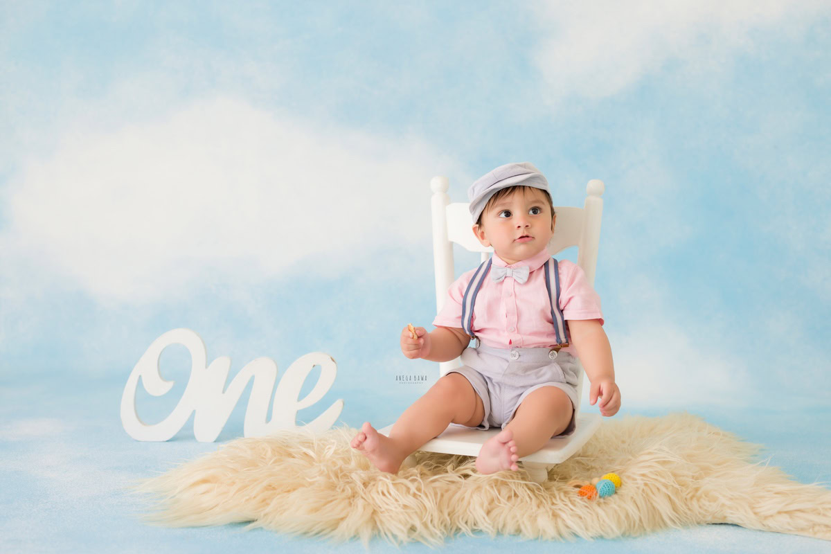 For an enchanting toddler photoshoot in Delhi, Gurgaon, and Noida by Anega Bawa, we present a delightful 1-year-old boy sitting on a white chair placed on a beige rug against a whimsical cloudy backdrop. This setup perfectly captures the innocence and charm of early childhood, making it a memorable session.