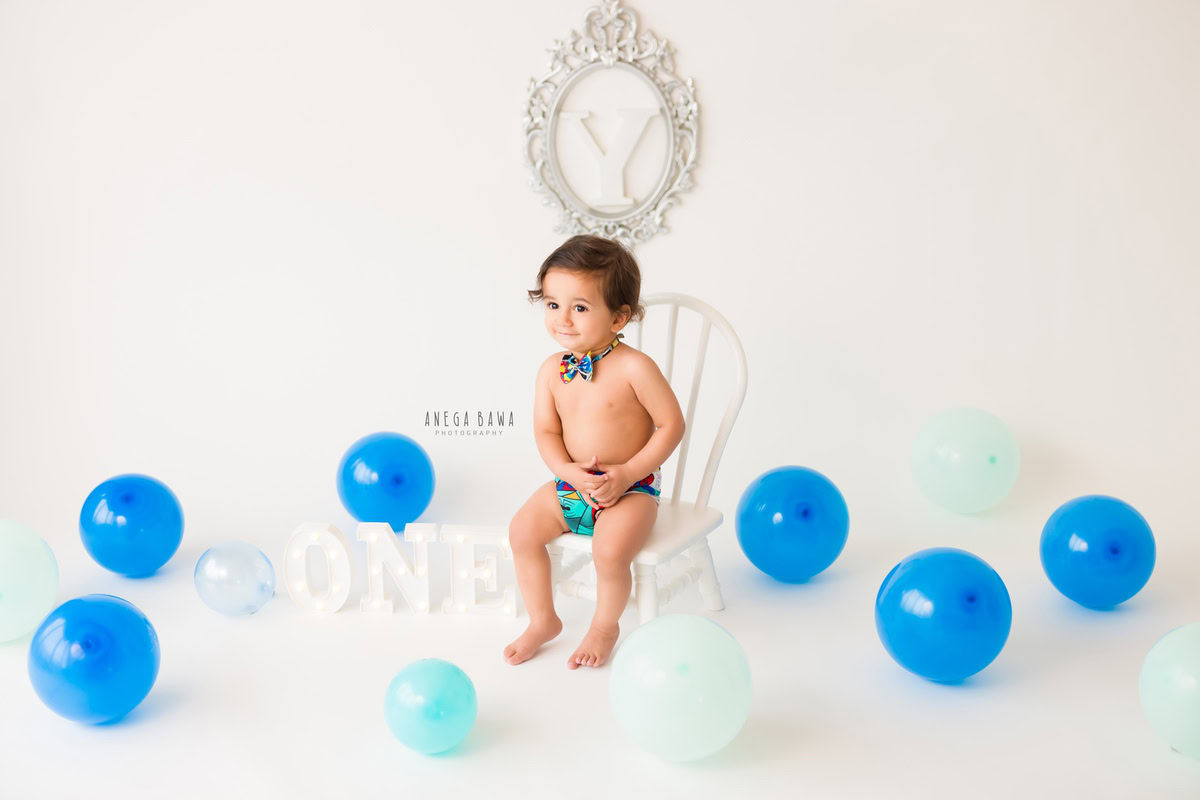 Adorable 1-year-old boy sitting on a charming white chair, pristine white backdrop, vibrant blue balloons, elegant decorative frame on the wall, professional sitter photographer in Gurgaon, Anega Bawa, Delhi.
