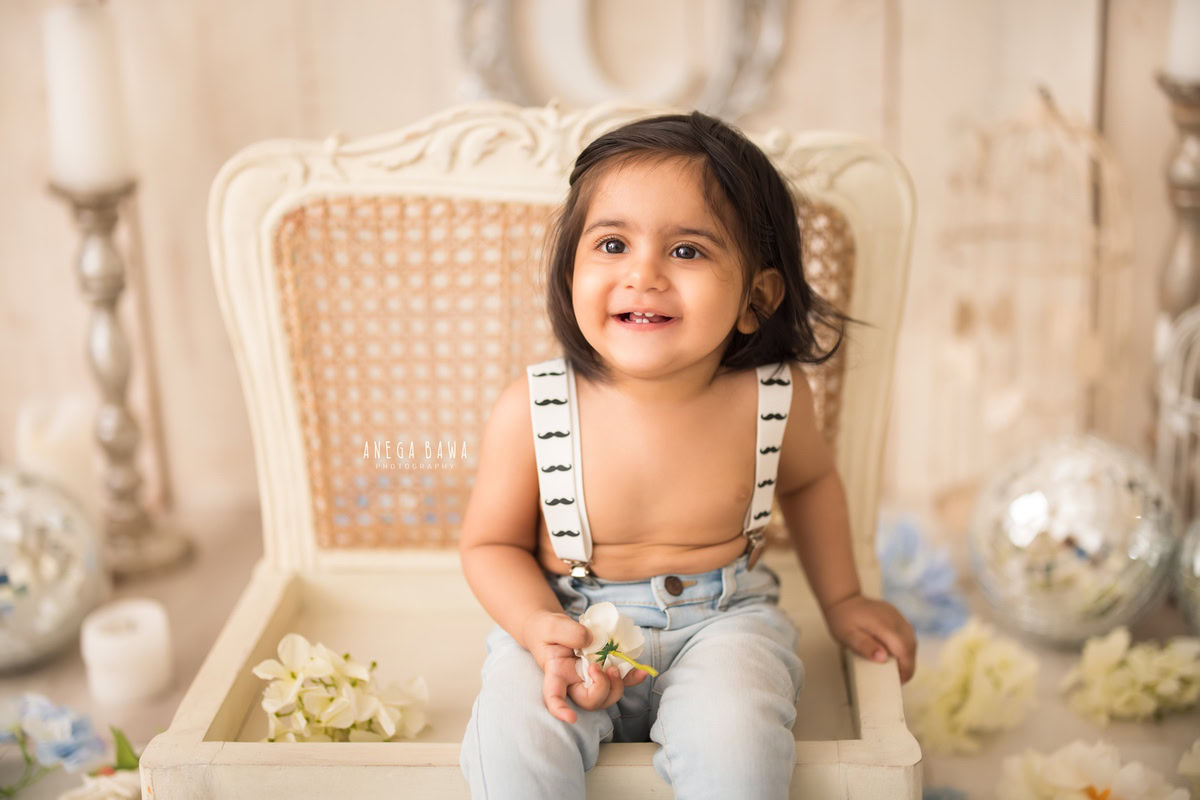 Capture the timeless innocence of a 1-year-old boy sitting on a white wooden chair, surrounded by a disco ball and candle stand against a beige backdrop. Perfect for a sitter photography session in Delhi and Gurgaon by Anega Bawa.