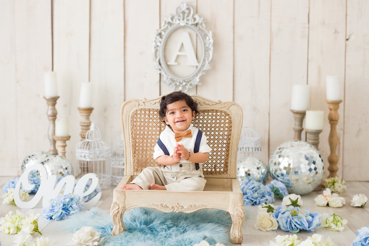 Capture the adorable innocence of your 1-year-old boy seated on a wooden chair, surrounded by the warm glow of candlelight, shimmering disco balls, and a playful blue rug adorned with blue pompoms. Trust Anega Bawa Photography in Delhi and Gurgaon to preserve these delightful moments with their expert sitter photography services.
