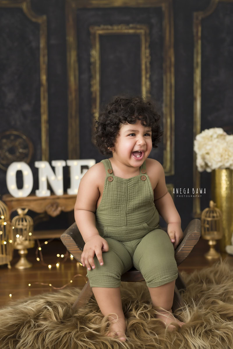 For an enchanting toddler photoshoot in Delhi, Gurgaon, and Noida by Anega Bawa, we present a delightful scene featuring a 1-year-old boy sitting on a wooden curvy chair with golden castles in the background. The setting is completed with a brown rug and fairy lights, capturing the magic and innocence of early childhood.
