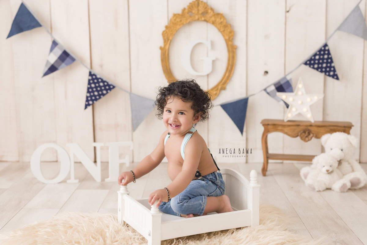 For a charming toddler photoshoot in Delhi, Gurgaon, and Noida by Anega Bawa, imagine a 1-year-old boy sitting in a white cot. The backdrop features an alphabet frame on the wall and adorable teddy bears against a beige background, creating a warm and inviting scene.