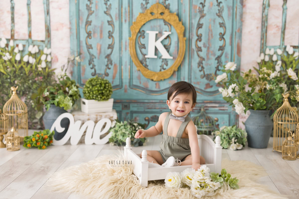 For a delightful toddler photoshoot in Delhi, Gurgaon, and Noida by Anega Bawa, picture a 1-year-old boy sitting in a white cot on a beige rug. The backdrop features golden castles and blue pots, with an alphabet frame on the wall set against a serene blue background.