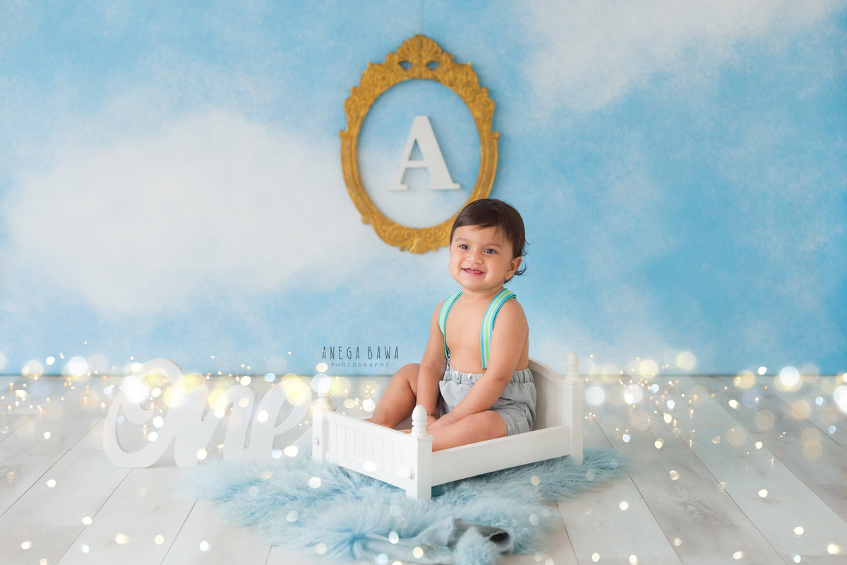 For a charming toddler photoshoot in Delhi, Gurgaon, and Noida by Anega Bawa, visualize a 1-year-old boy sitting in a white cot on a blue rug. The backdrop is adorned with fairy lights, an alphabet frame on the wall, all set against a soothing blue background.