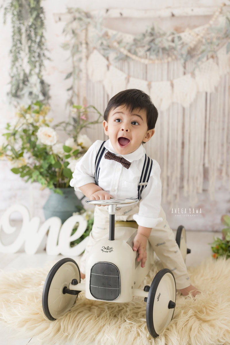 Imagine capturing delightful moments of a 1-year-old boy during a toddler photoshoot in Delhi, Gurgaon, and Noida by Anega Bawa. Picture him seated on a toy scooter against a backdrop of lush green pots, all set on a clean white backdrop.