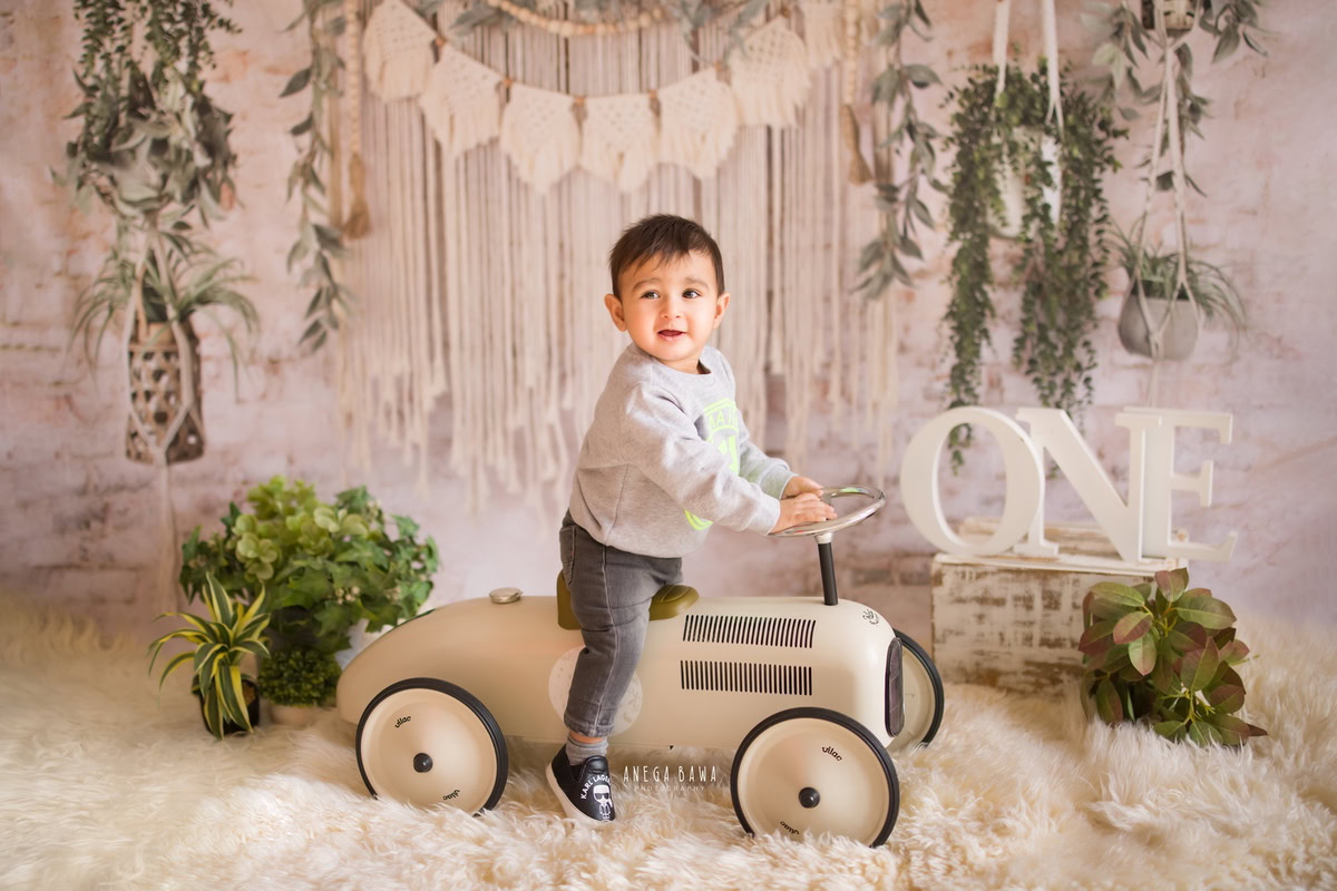 Capture the joy of a 1-year-old boy sitting on a toy scooter, set against a white backdrop with hanging leaves and green pots. Ideal for a sitter photography session in Delhi and Gurgaon by Anega Bawa.
